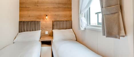 A double room with two beds, a wooden headboard, a nightstand, a wall-mounted light, and a window with curtains.