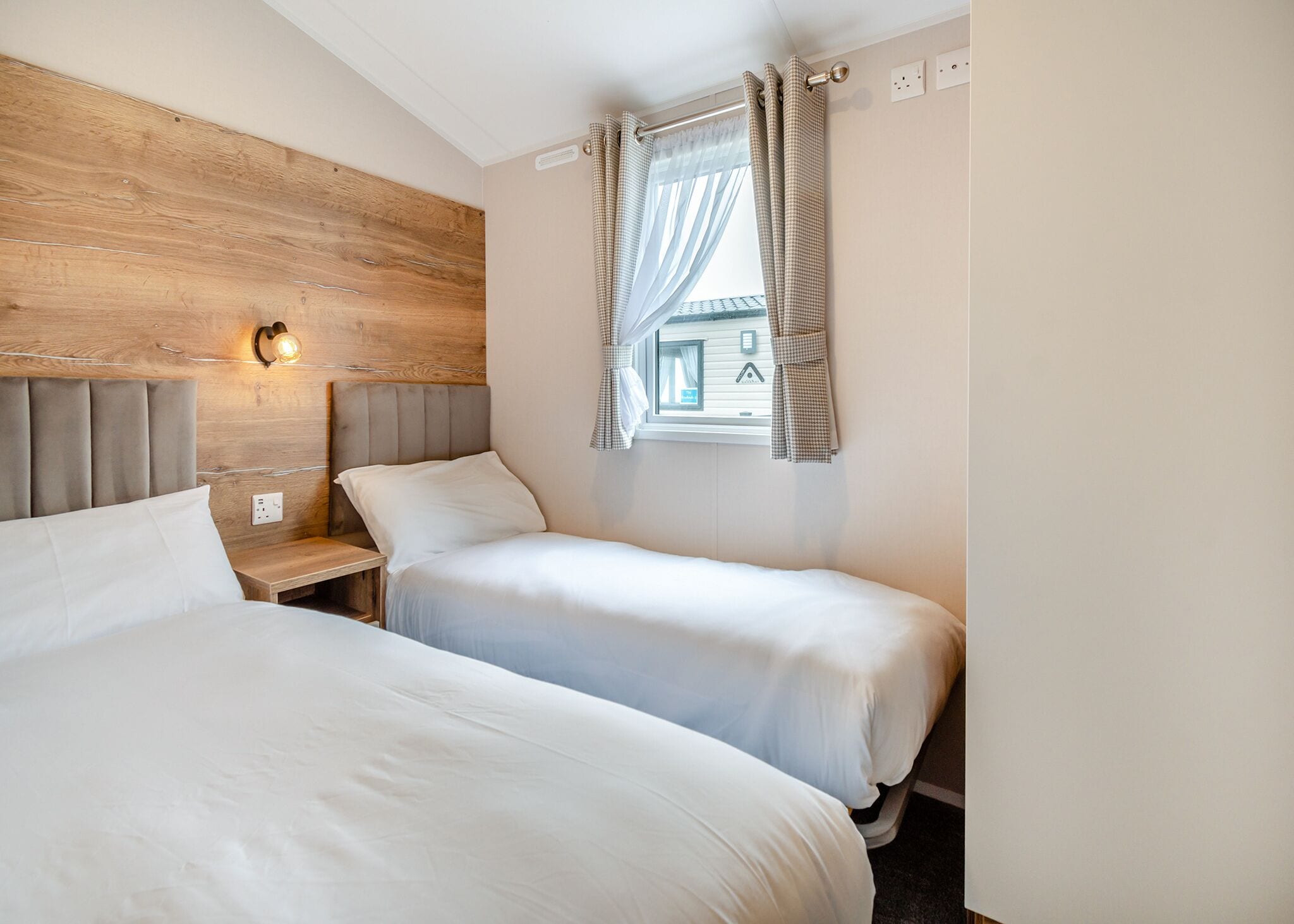 A bedroom with two beds, a wooden headboard, a nightstand with a lamp, and a window with curtains.