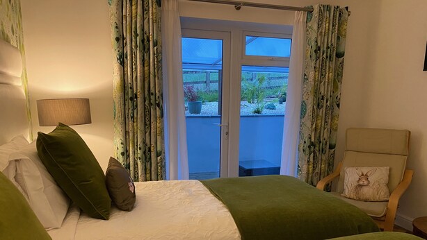 A hotel room with two beds, a chair with a pillow, a lamp, and a view of a balcony with plants. - Toppesfield House (Bude)