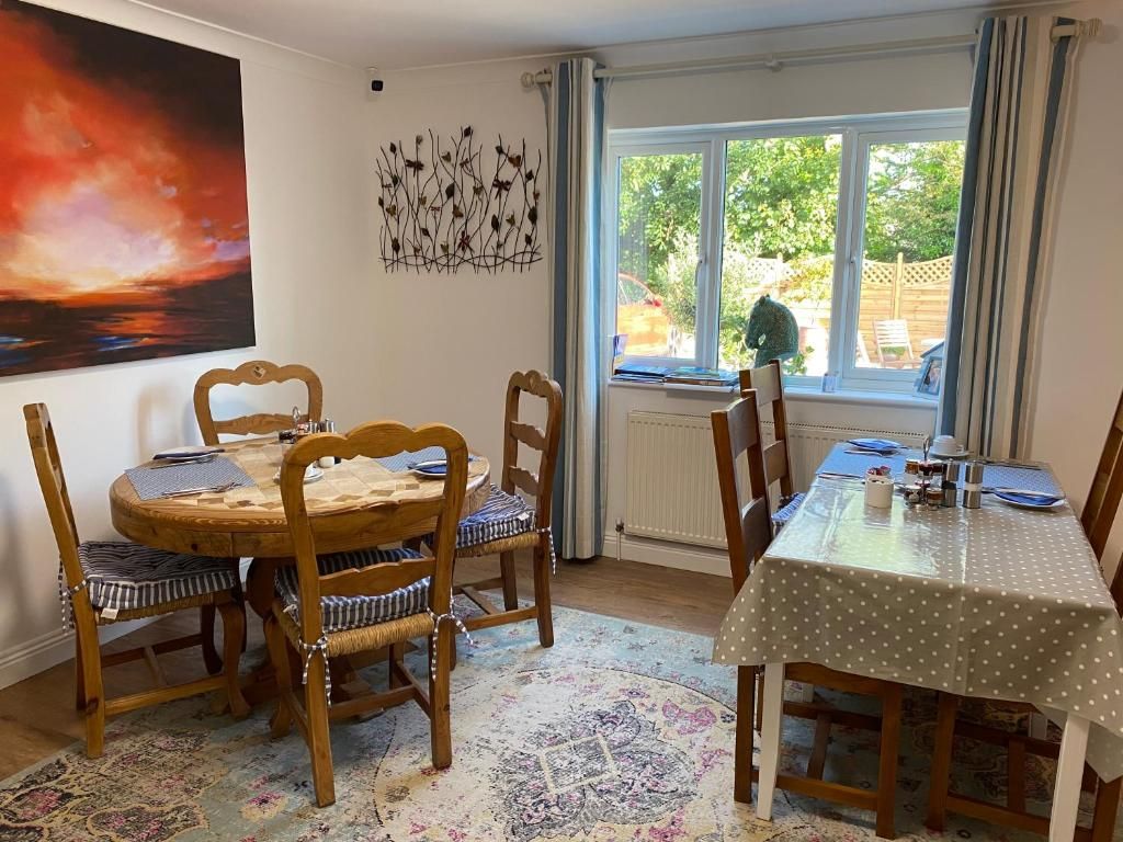 A dining area with two wooden tables, one with a polka dot tablecloth and another with a placemat. There are four chairs with striped cushions. A large painting is on the wall, and a window with curtains is visible.