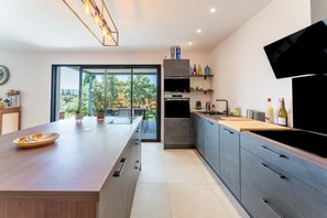 A modern kitchen with a central island, built-in appliances, and a view through glass doors. - Villa 'Mas Mayflower' with Mountain View, Wi-Fi, and Air Conditioning (Cabrières-d'Avignon)