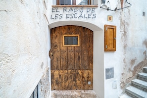 Una rustica porta di legno con un piccolo oblò, un cartello sopra con la scritta 'EL RACO DE LES ESCALES' e il numero '6' sul muro.