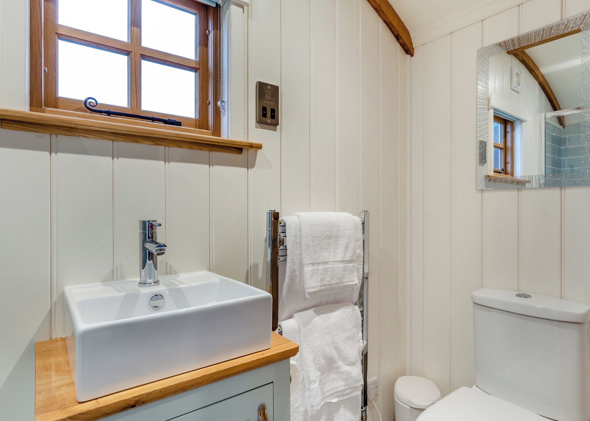 Un baño con lavamanos blanco, toallero, inodoro y ventana con marco de madera.