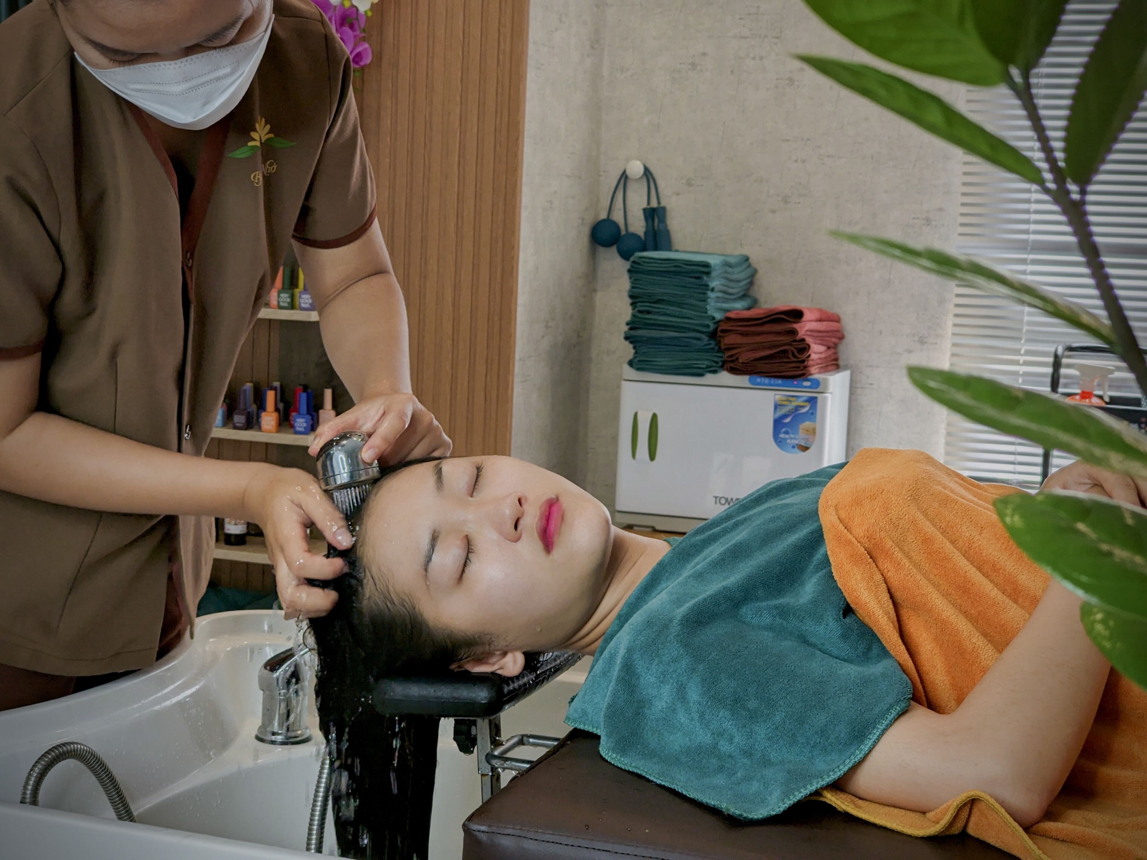 A person receiving a hair treatment in a bathroom with a sink, a towel, and a shelf with various items.