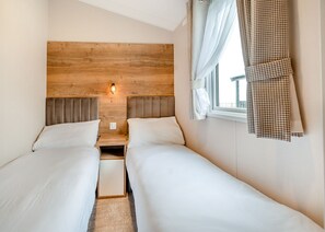 A double room with two beds, a wooden headboard, a nightstand, a wall-mounted light, and a window with curtains. - This stunning holiday home with a double bedroom and en suite W. (Hornsea)