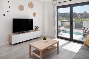 A modern living room with a flat-screen TV, a wooden coffee table, a potted plant, and a sliding glass door leading to a balcony with a pool.