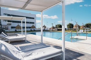 Un espace aménagé au bord de la piscine, avec des chaises longues, un coin salon abrité et un bâtiment moderne en arrière-plan.