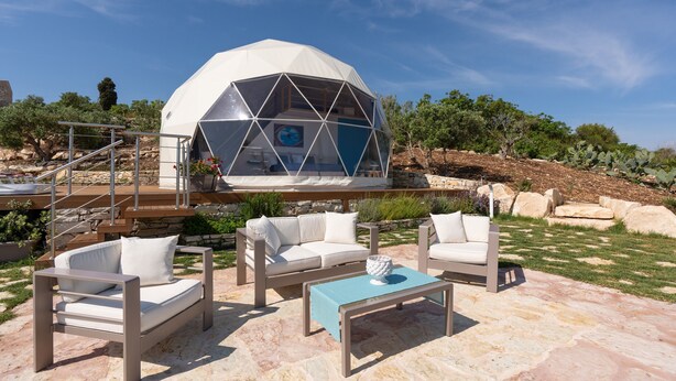 Una estructura de domo geodésico con una escalera que lleva a un nivel superior, rodeada de asientos exteriores con cojines blancos y una mesa azul.