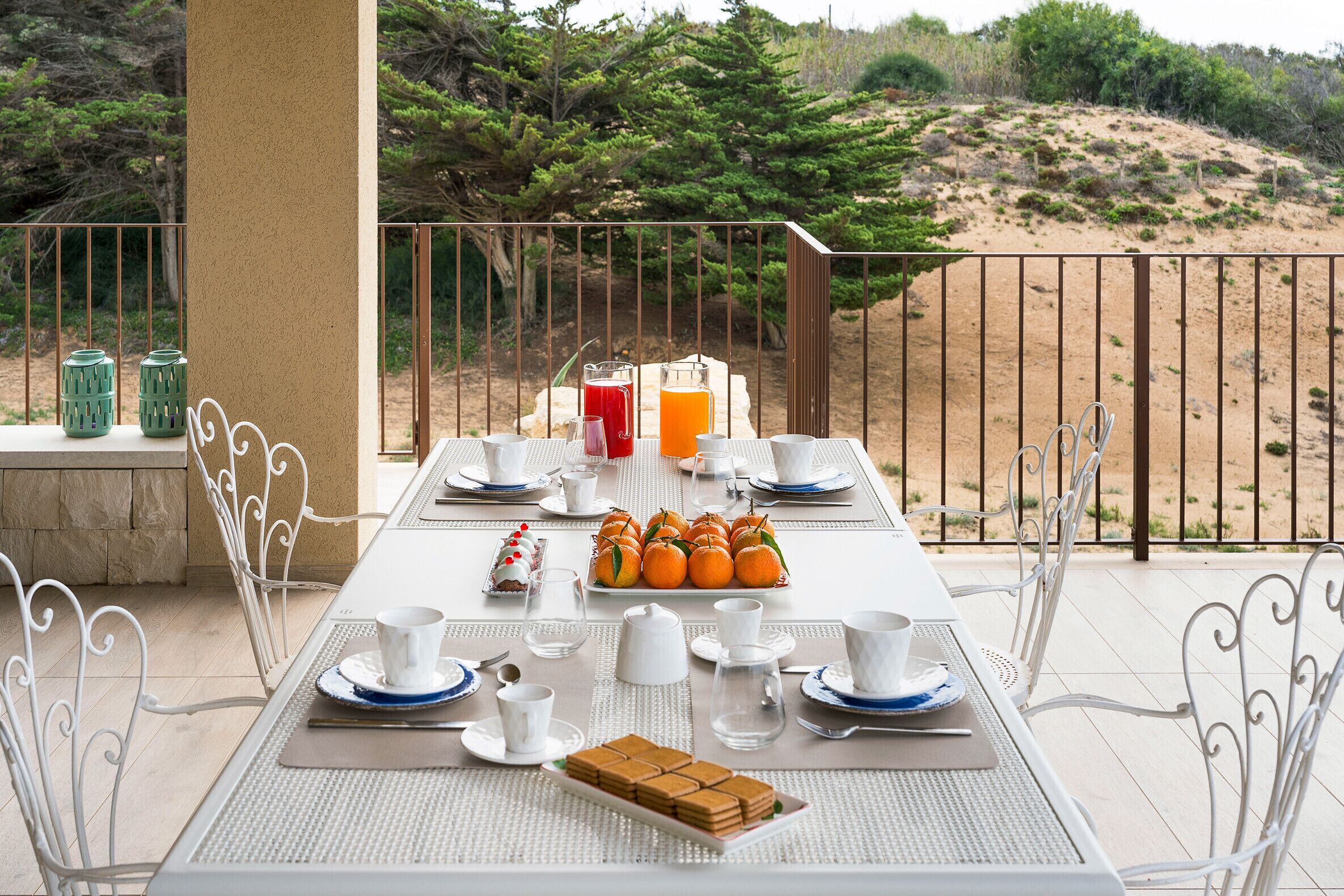 Une table à manger est dressée avec de l’argenterie blanche et bleue, des tasses et un plateau de fruits, et offre une vue sur un paysage sablonneux parsemé d’arbres.