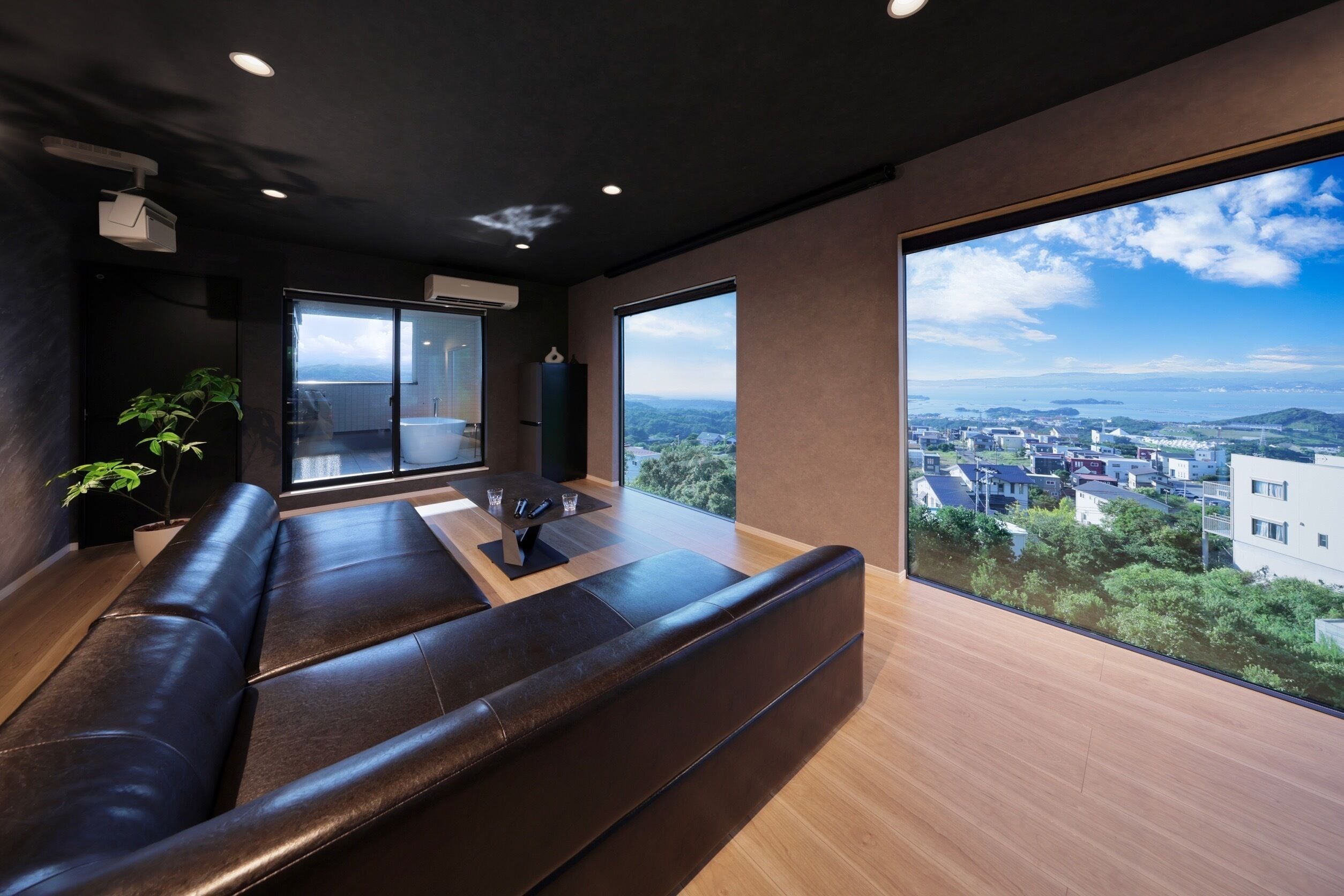 Ein modernes Wohnzimmer mit großem Fenster und Blick auf die Stadt, einem dunklen Ledersofa und einem kleinen Tisch.