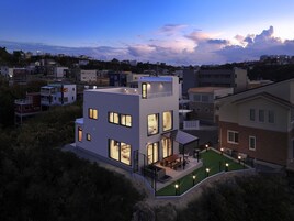 Una casa moderna de dos pisos con techo plano, ventanas amplias y un pequeño patio con mobiliario exterior.