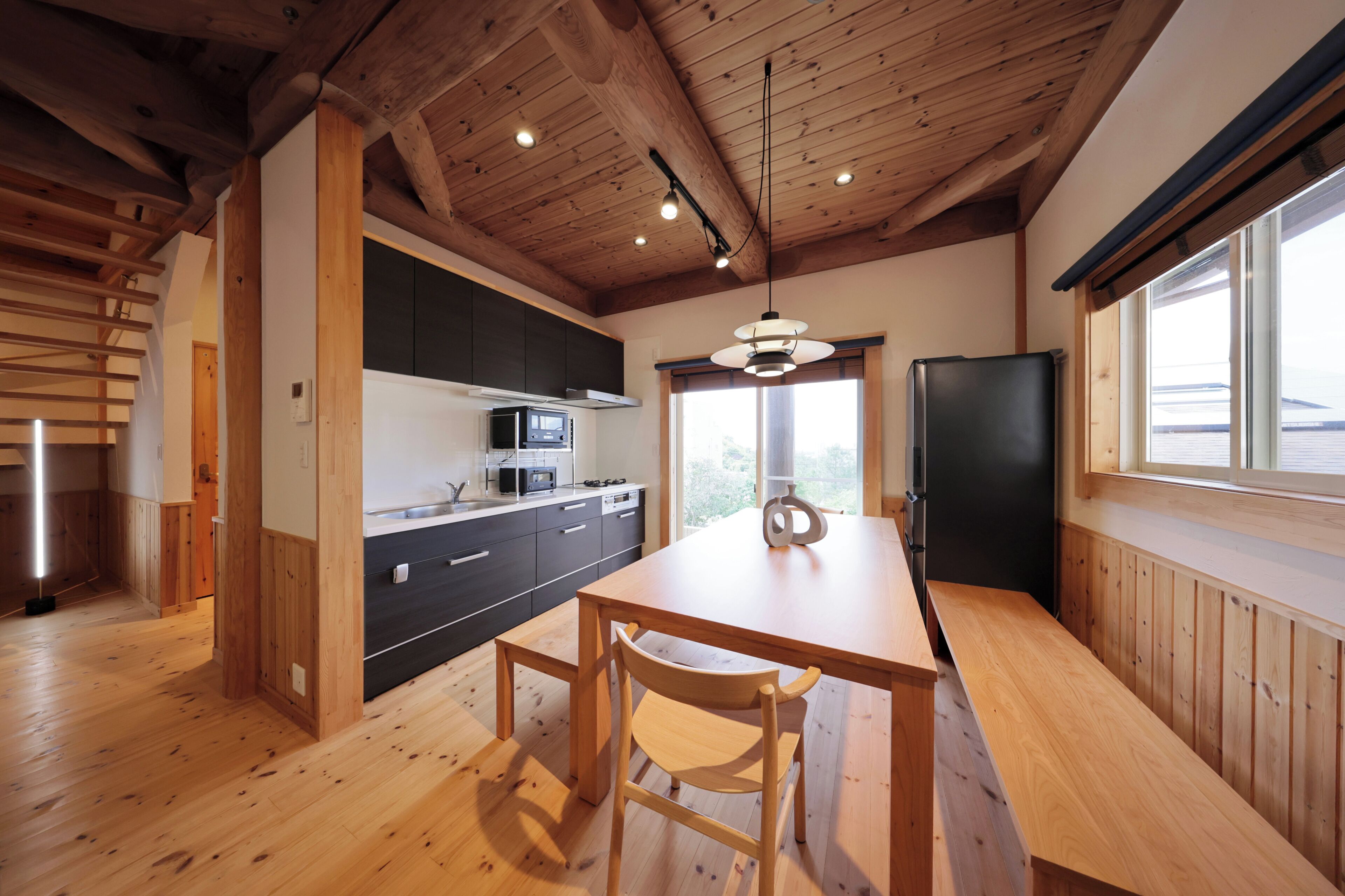 A kitchen with wooden flooring, a dining table with chairs, a black refrigerator, and a window with a view.