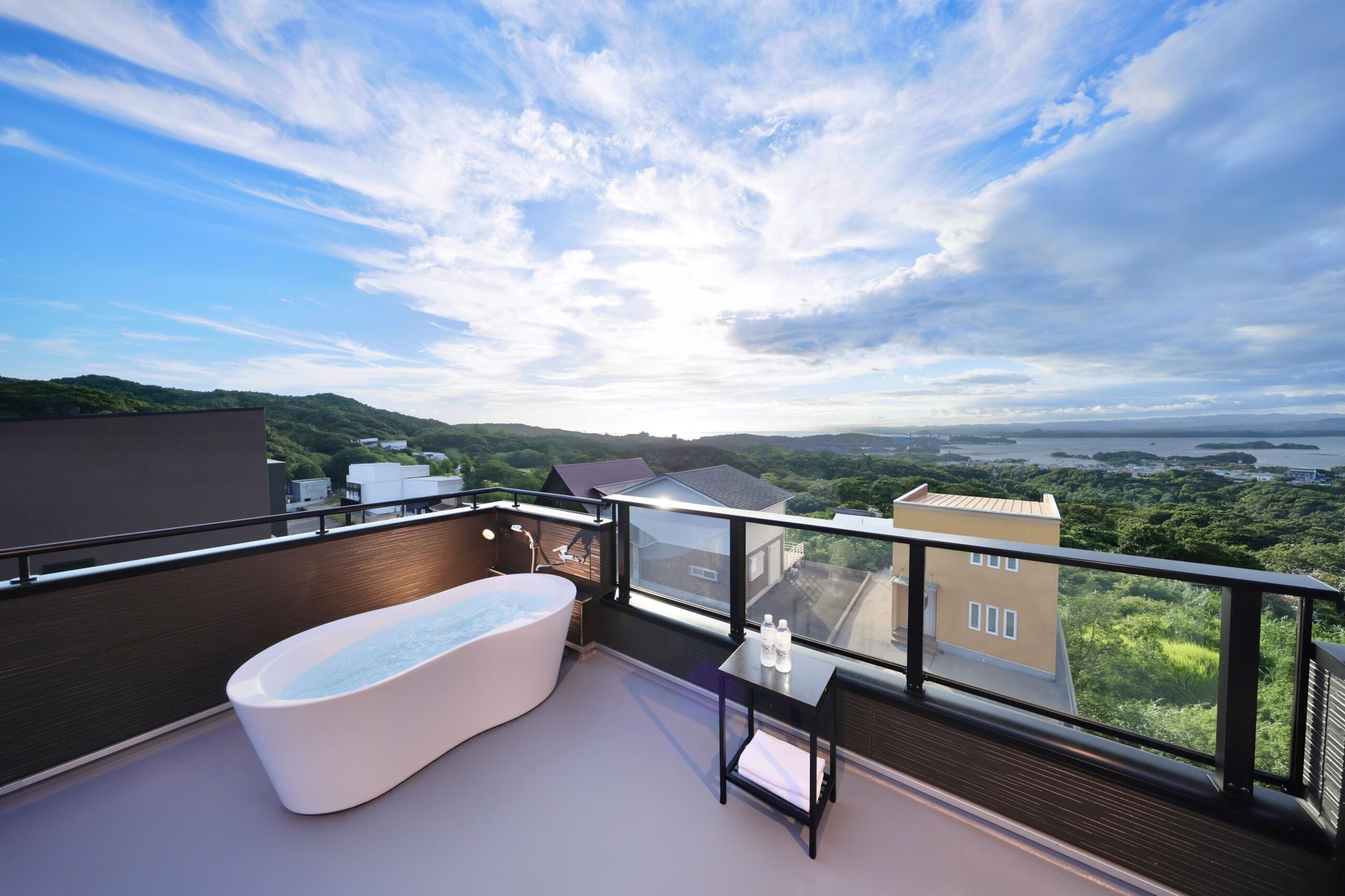 A balcony with a bathtub, a table with bottles, and a view of a green landscape and buildings.