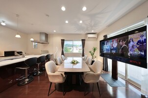 Un comedor moderno con una mesa grande, taburetes de bar y un televisor de pantalla plana mostrando una película.