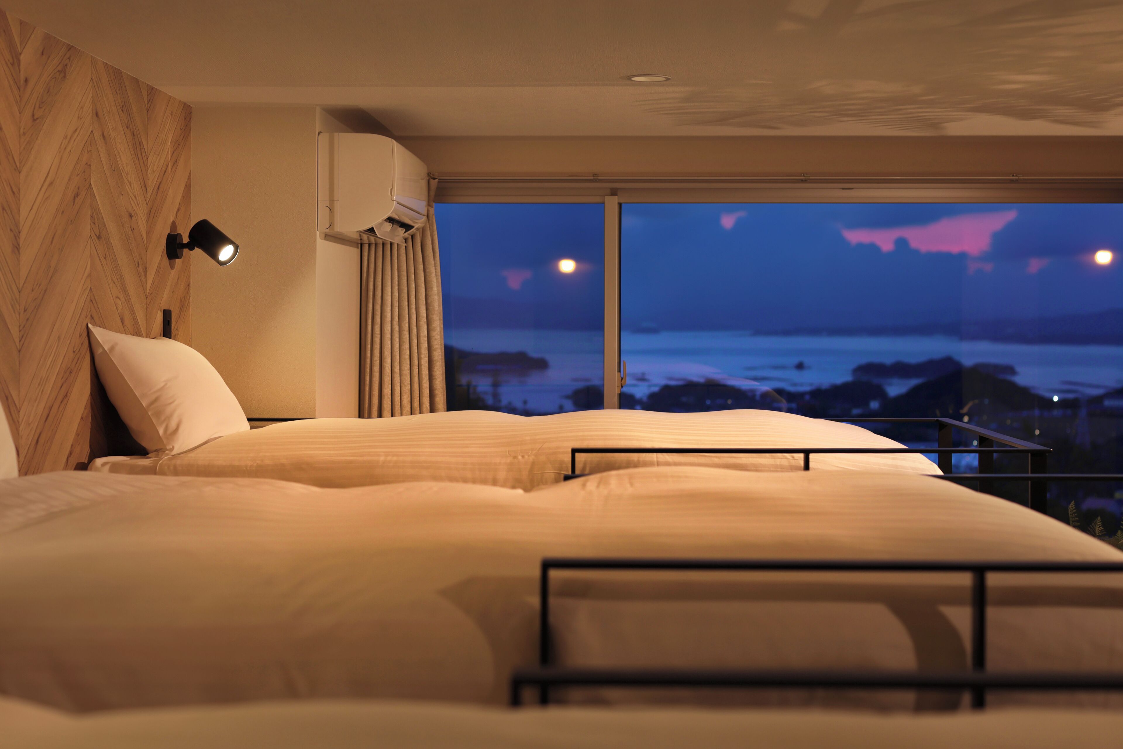 Une chambre avec un grand lit, une table de chevet, une lampe et une vue sur l’océan au crépuscule.
