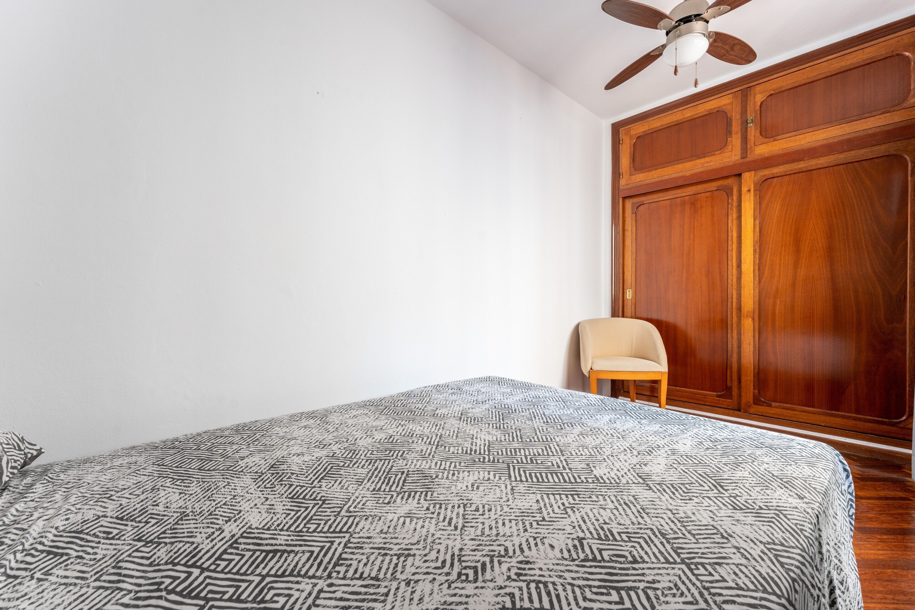 Un dormitorio con cama, ventilador de techo, armario de madera y una silla.