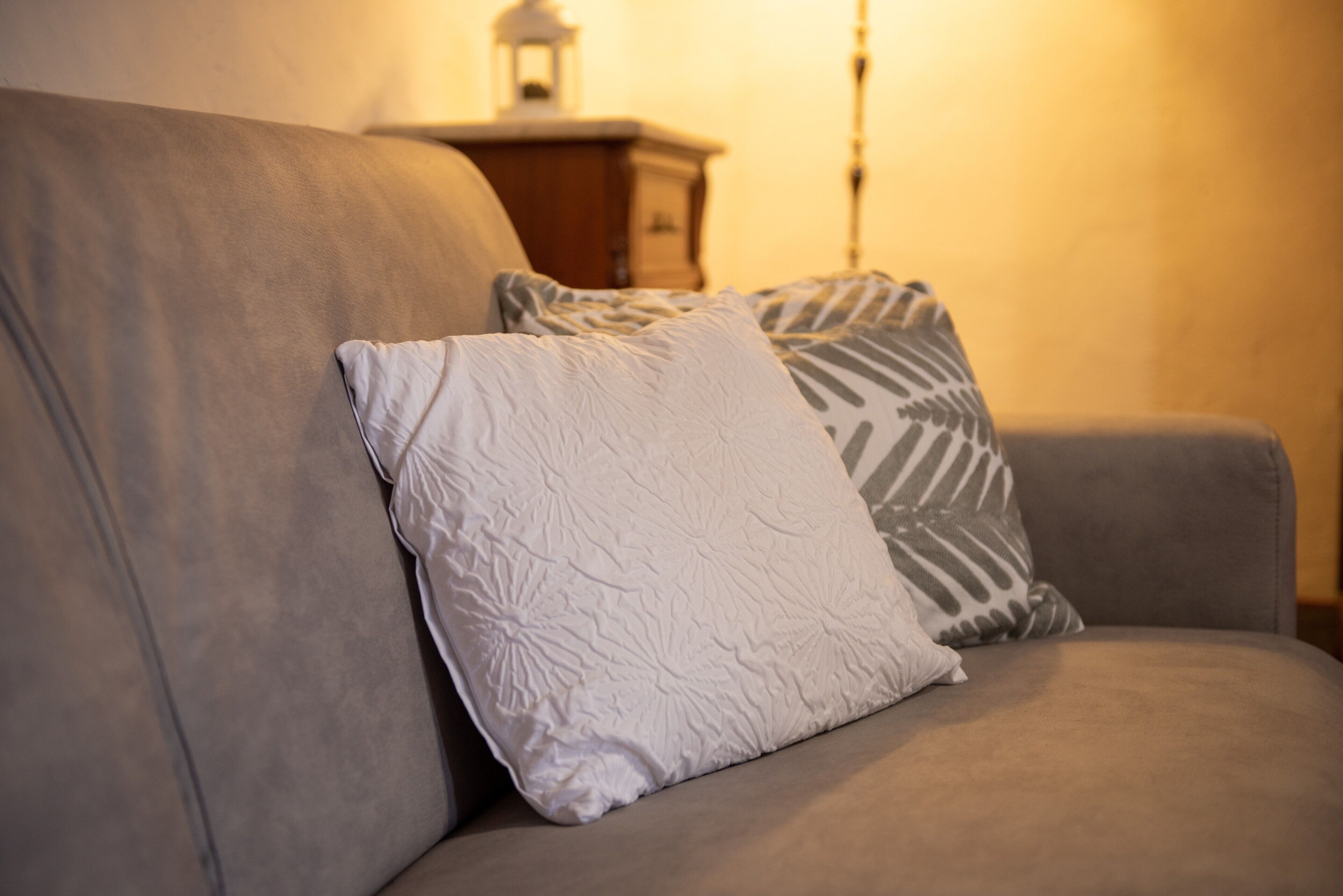 A sofa with two pillows, a wooden side table with a lamp, and a wall with a textured pattern.