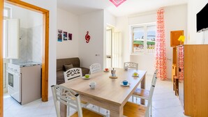 Een eetgedeelte met een houten tafel, witte stoelen, een kleine keuken met een fornuis en een roze hanglamp.