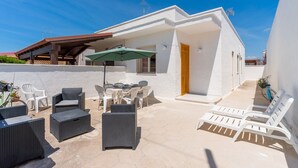 Een witte villa met buitenmeubilair, waaronder rieten stoelen en een tafel, en een fiets tegen de muur geleund.