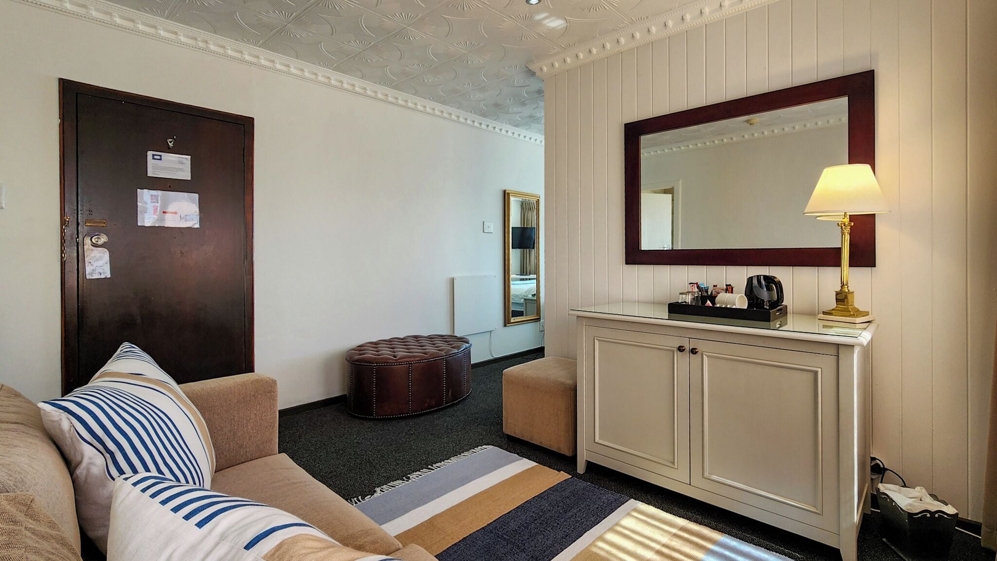Et hotelværelse med en sofa, skærm, et lille bord med lampe, et spejl og et skab.