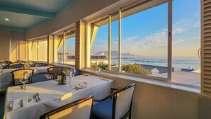 A dining area with tables set for a meal, large windows offering a view of the sea and distant buildings. - Blue Peter Hotel (Cape Town)