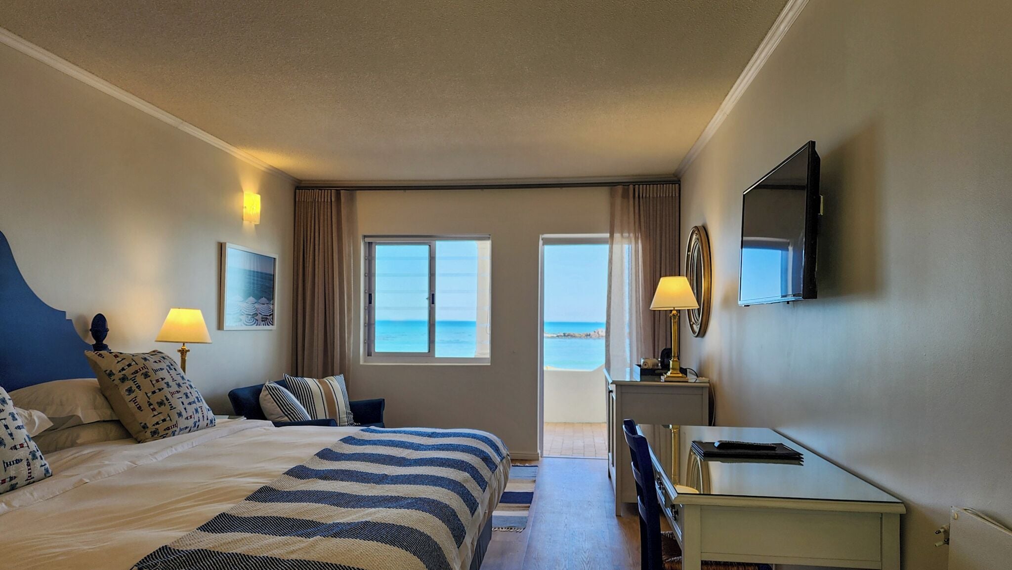 A hotel room with a bed, a desk, a TV, and a view of the sea through the windows.
