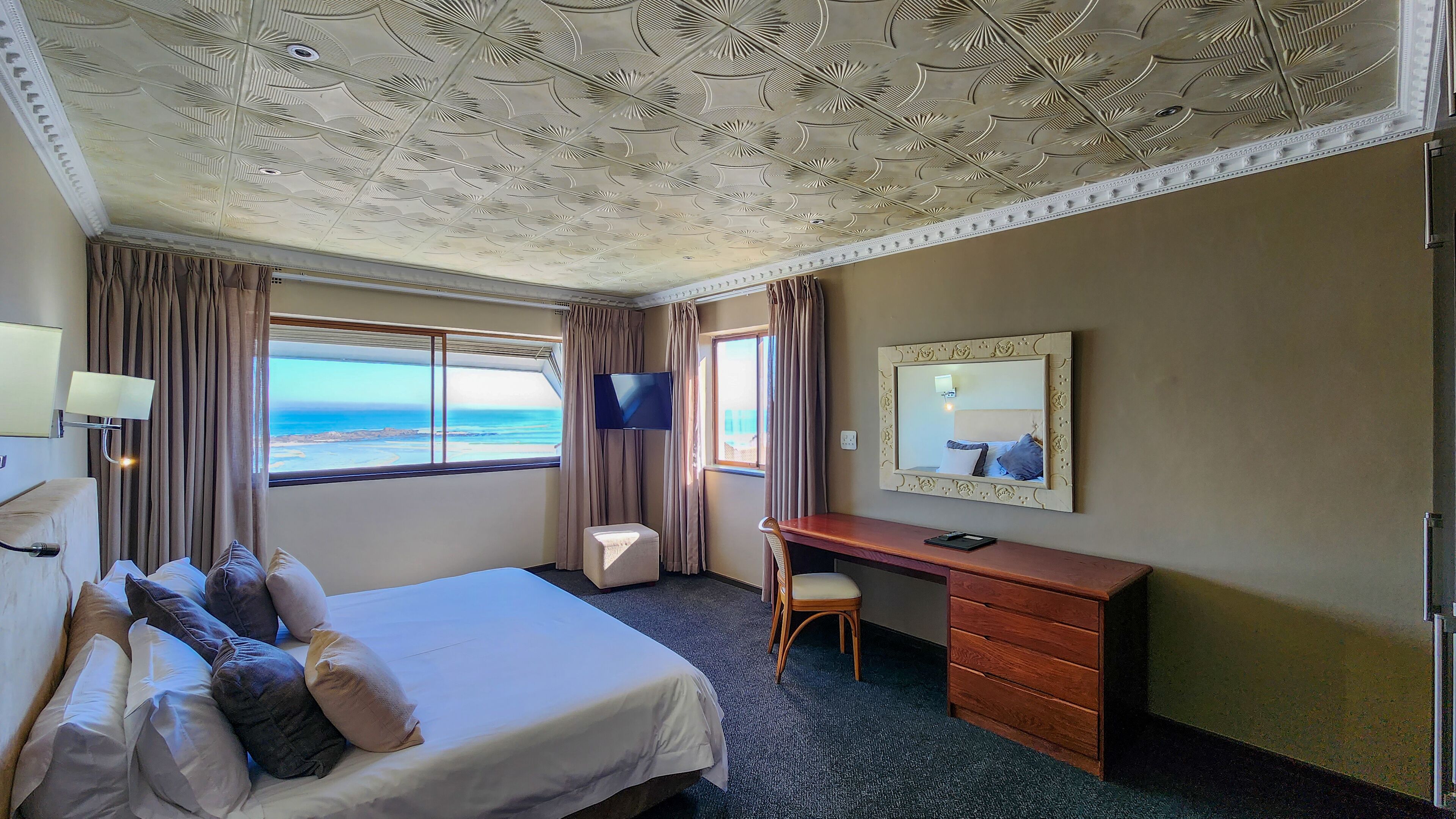 A hotel room with a large bed, a desk, a chair, a TV, and a window with a view of the sea.