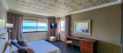 A hotel room with a large bed, a desk, a chair, a TV, and a window with a view of the sea.