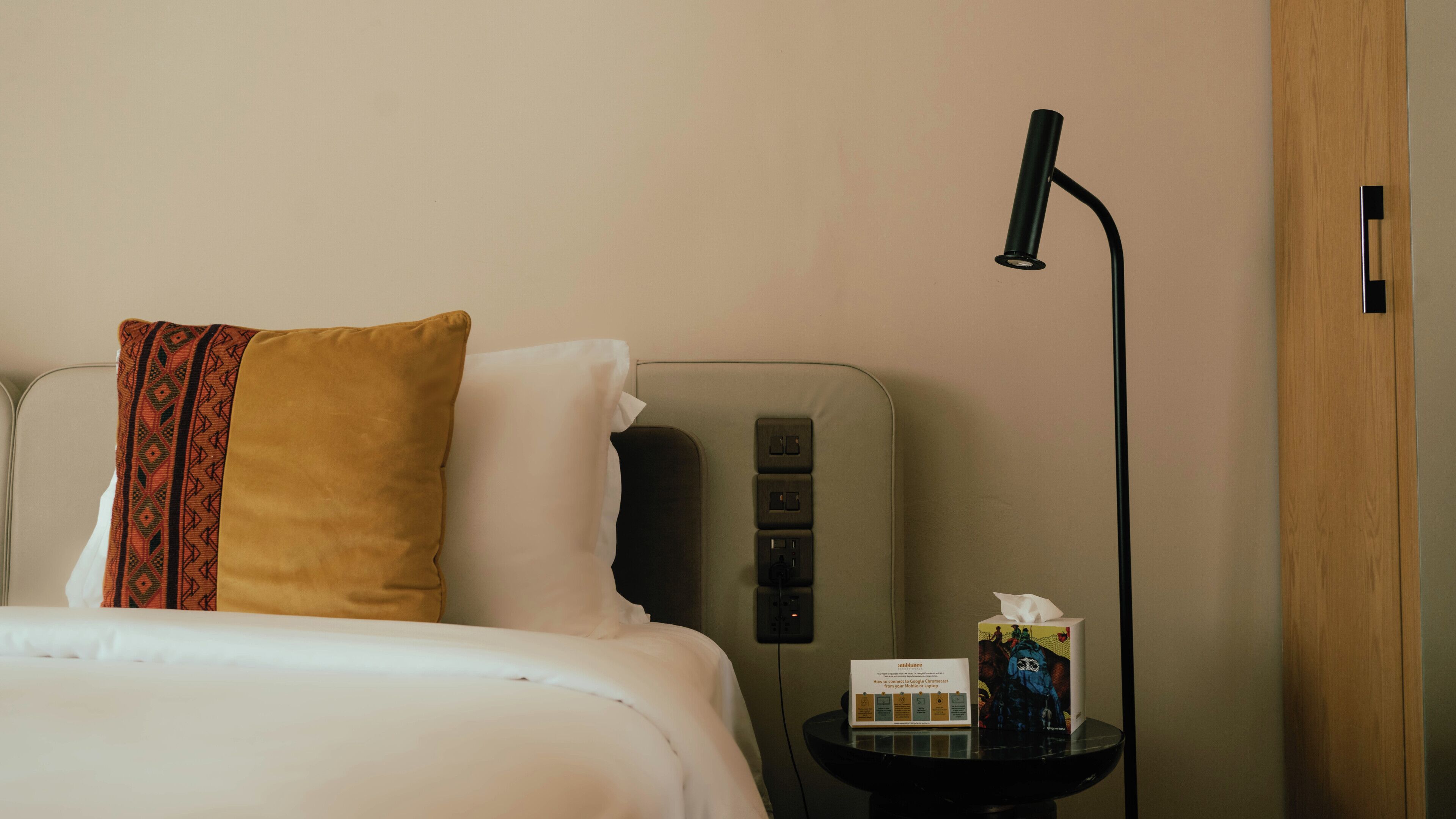 A hotel room with a bed, a bedside table with a tissue box and a booklet, a wall socket, and a floor lamp.