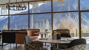 A restaurant with a mountain view, featuring a round table with chairs, a bar area, and a large window with a view of snow-capped mountains. - Ambiance Resort Hunza (Karimabad)