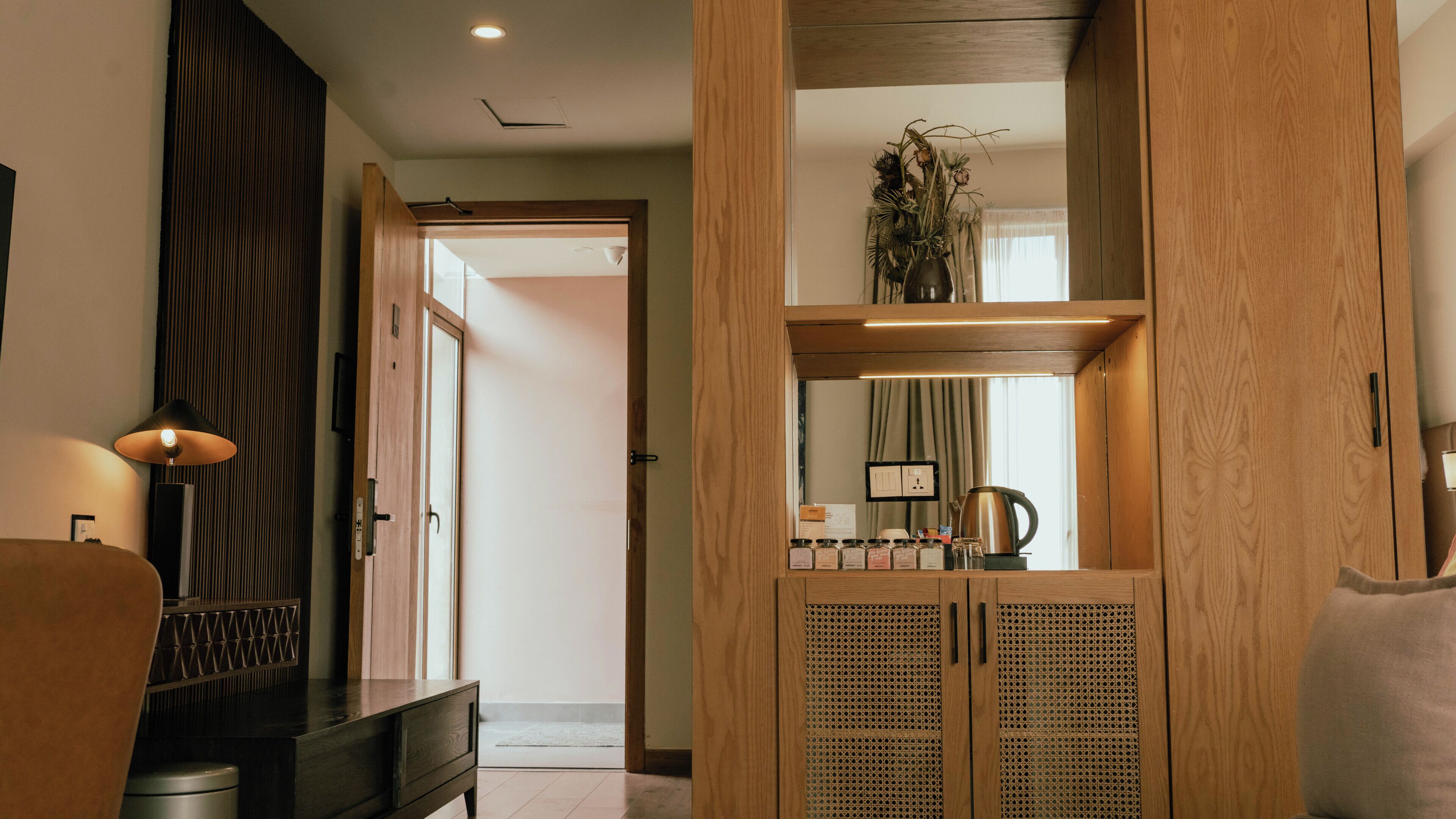 A hotel room with a wooden wardrobe, a desk, a lamp, and a mirror.