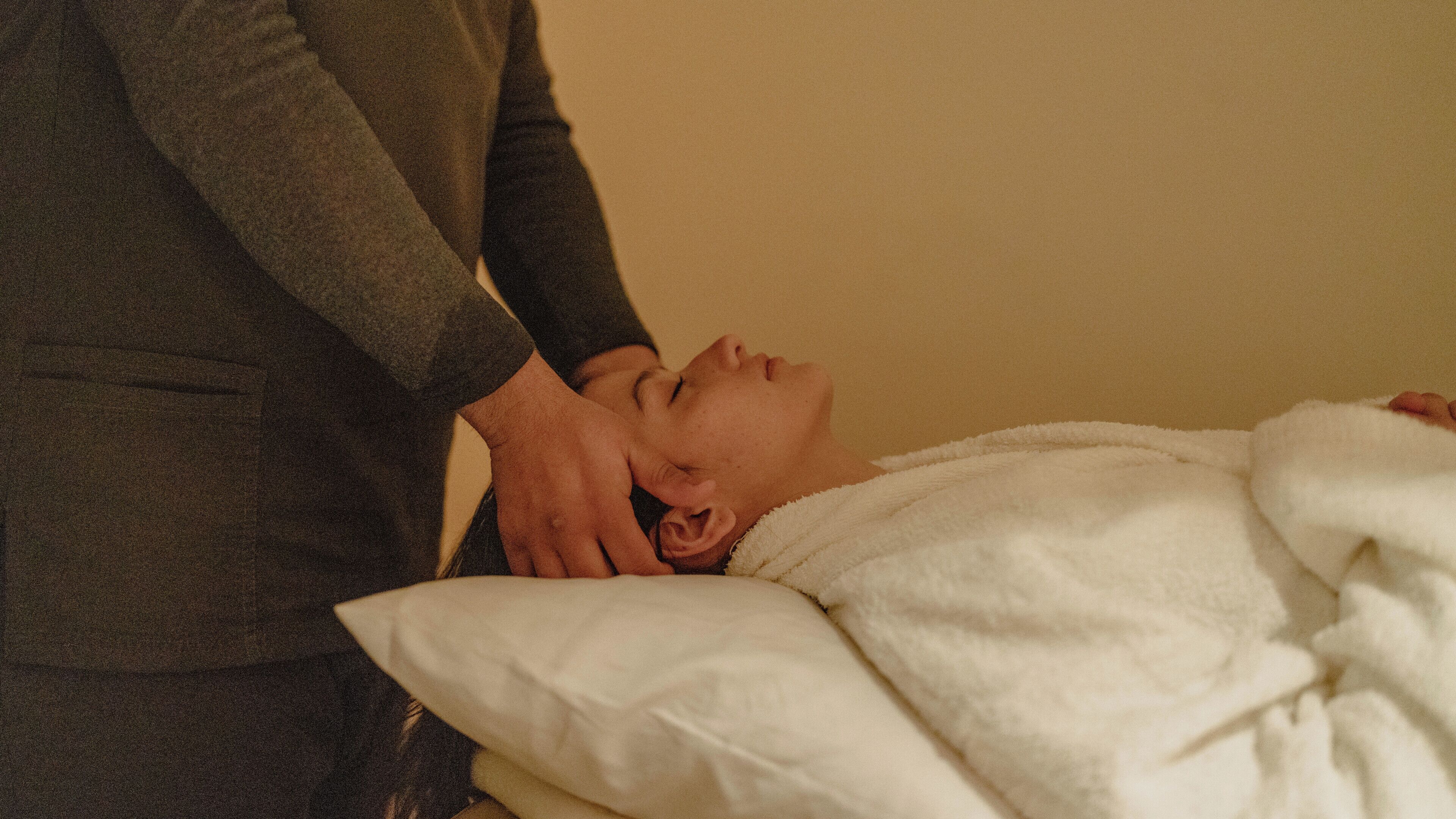 A person lying on a bed with a white blanket, receiving a massage from another individual.