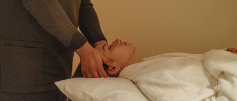 A person lying on a bed with a white blanket, receiving a massage from another individual.