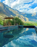 Una piscina cristalina con vistas a montañas, una terraza de madera con sillas y una zona de estar cubierta.