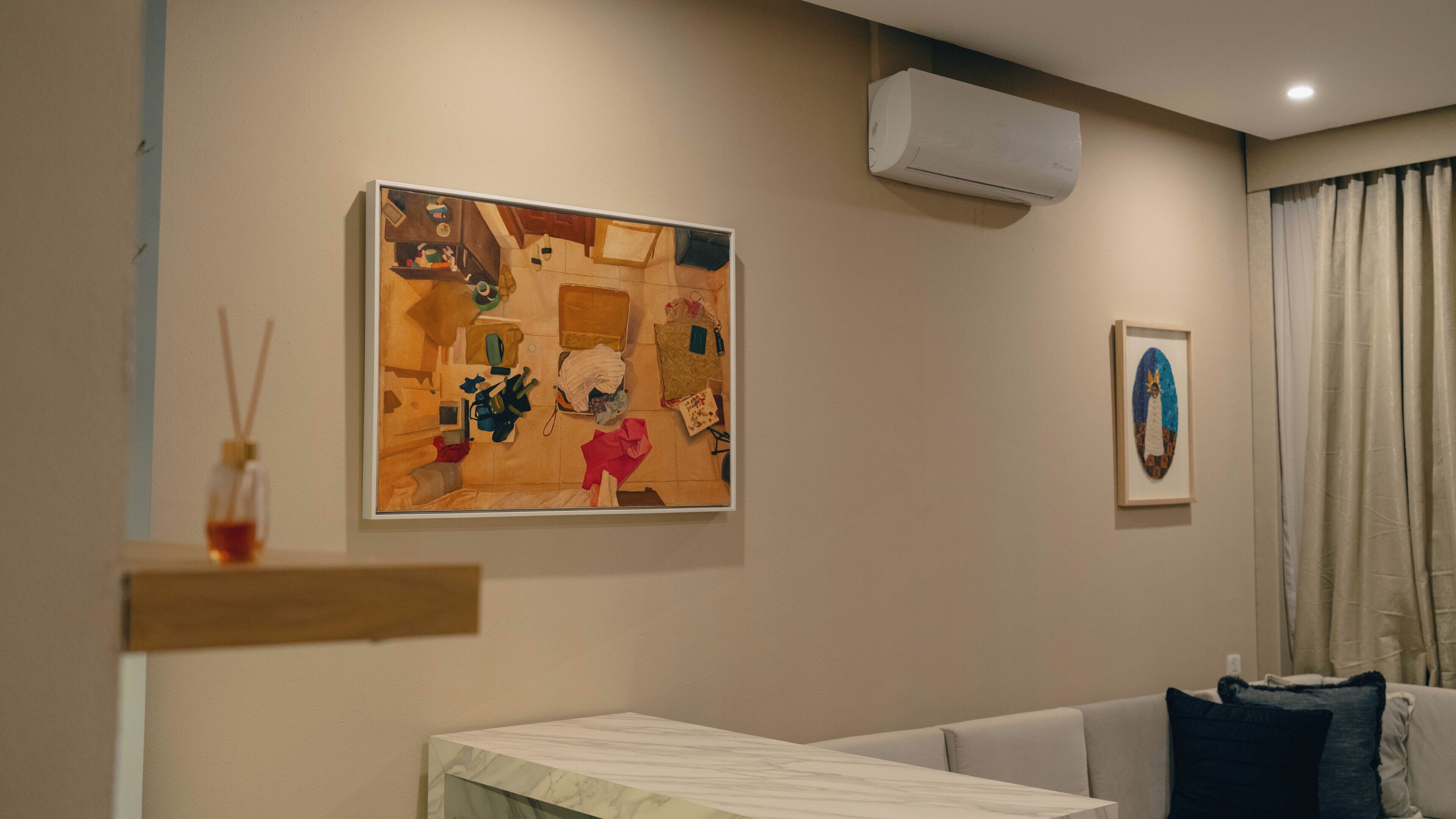 A room with a bed, a wall-mounted air conditioner, a framed abstract painting, and a small table with a bottle of perfume.