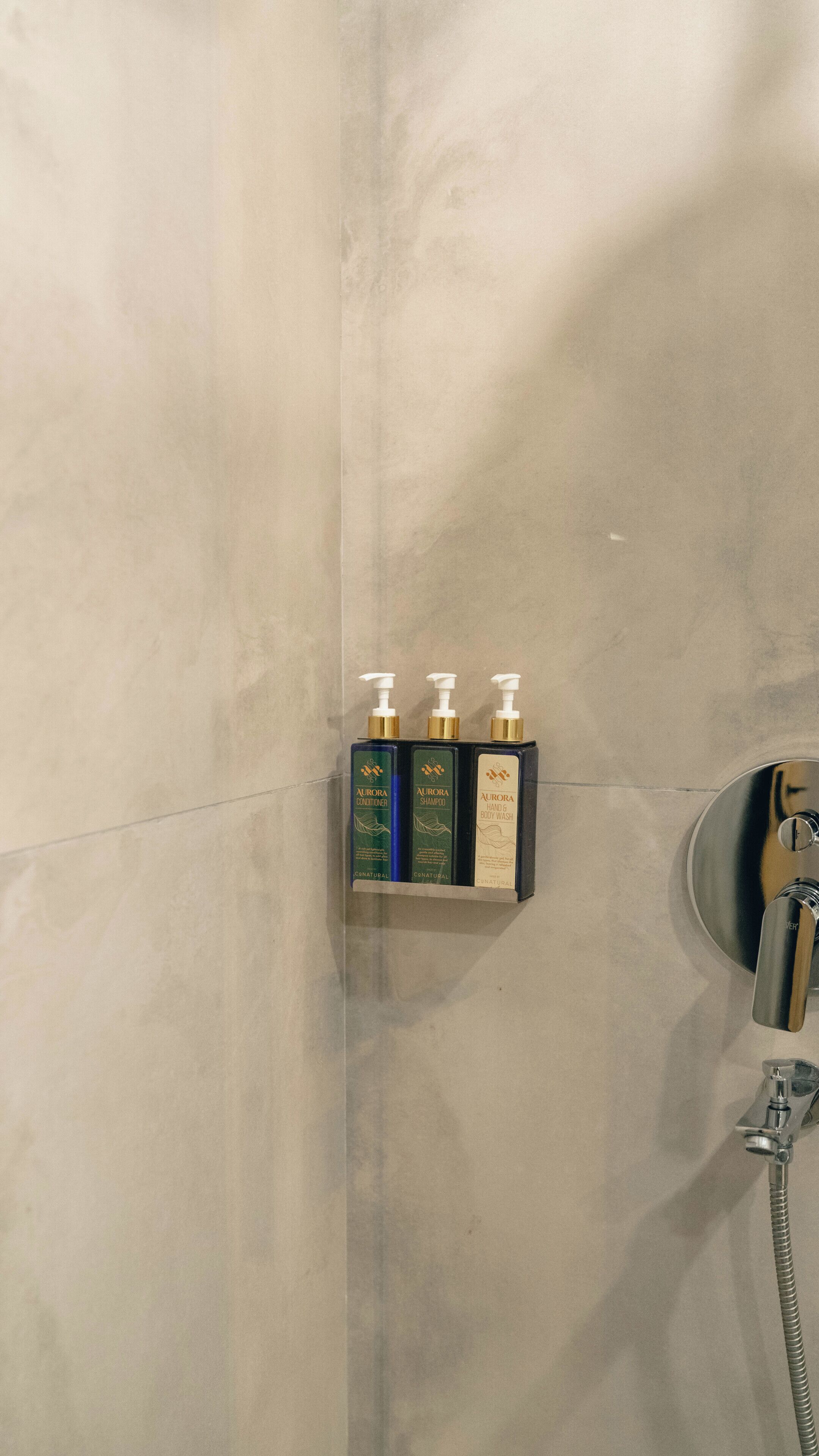 Un baño con ducha, un secador de manos y tres botellas de productos Aurora.