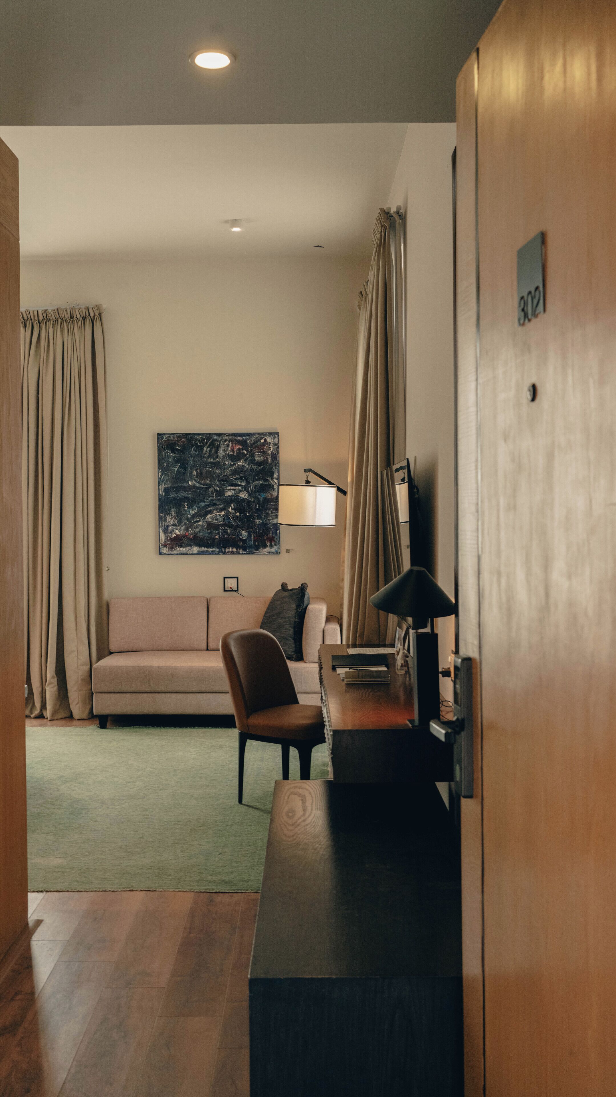 A modern hotel room with a wooden desk, a sofa, a chair, a painting on the wall, and a door with a number.