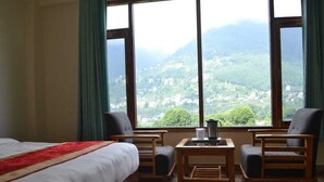A hotel room with a bed, two armchairs, a small table, and a large window offering a view of a mountainous landscape.