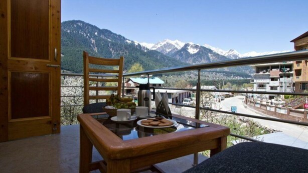 A balcony with a wooden table set for breakfast, a chair, and a view of mountains and buildings.