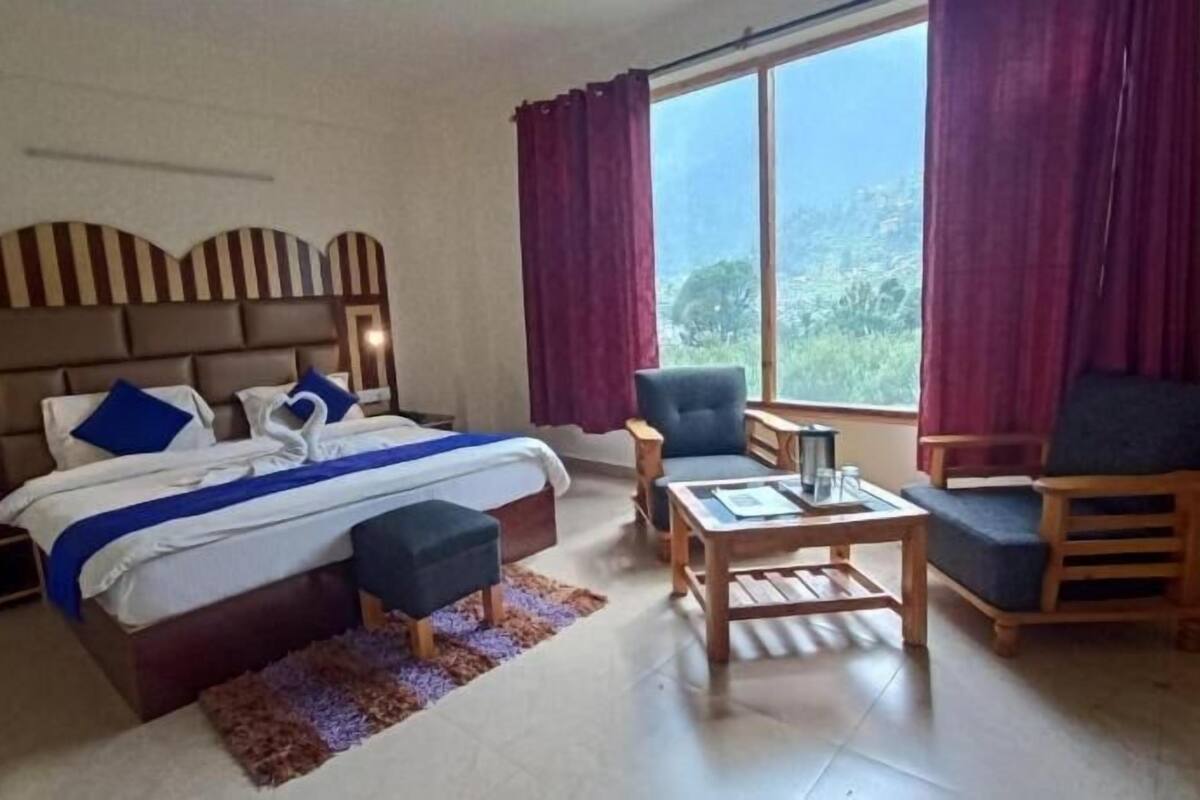 A hotel room with a bed, a desk, two chairs, a window with curtains, and a view of the outdoors.