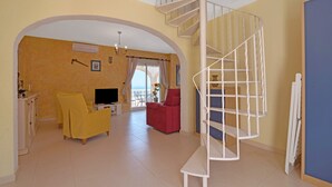 Un salon avec un canapé jaune, un fauteuil rouge, un téléviseur et un escalier en colimaçon.