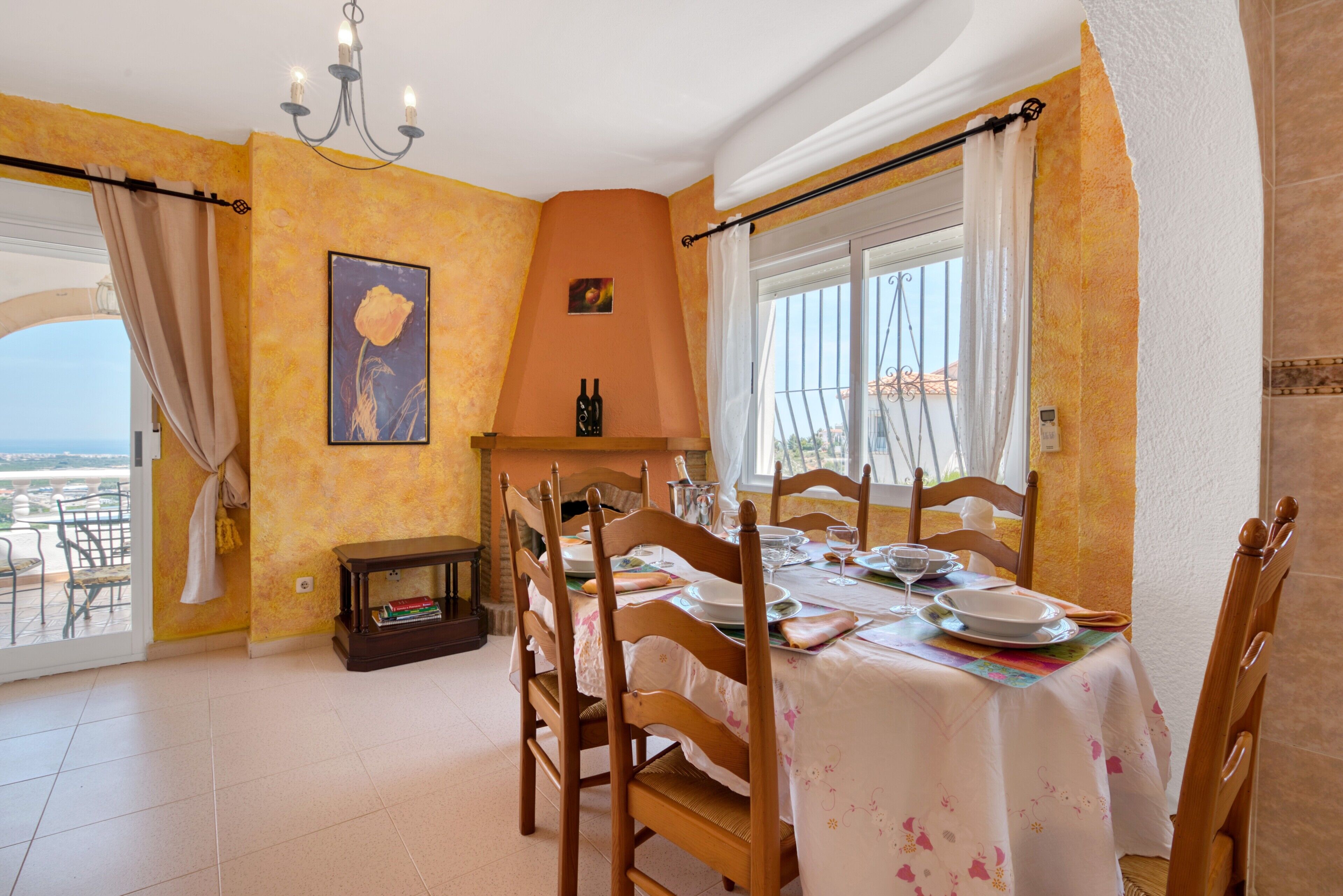 A dining room with a table set for four, wooden chairs, a chandelier, a painting, and a view through a window.