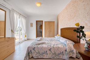 Ein Schlafzimmer mit einem Bett, einer Holzkommode, einem Nachttisch und einem Fenster mit Vorhängen.