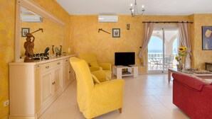 Un salon avec un fauteuil jaune, une télévision, un canapé rouge et un balcon avec vue.