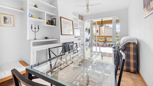 Une pièce moderne avec une table à manger en verre, des étagères blanches, un ventilateur de plafond et une vue sur l’extérieur.