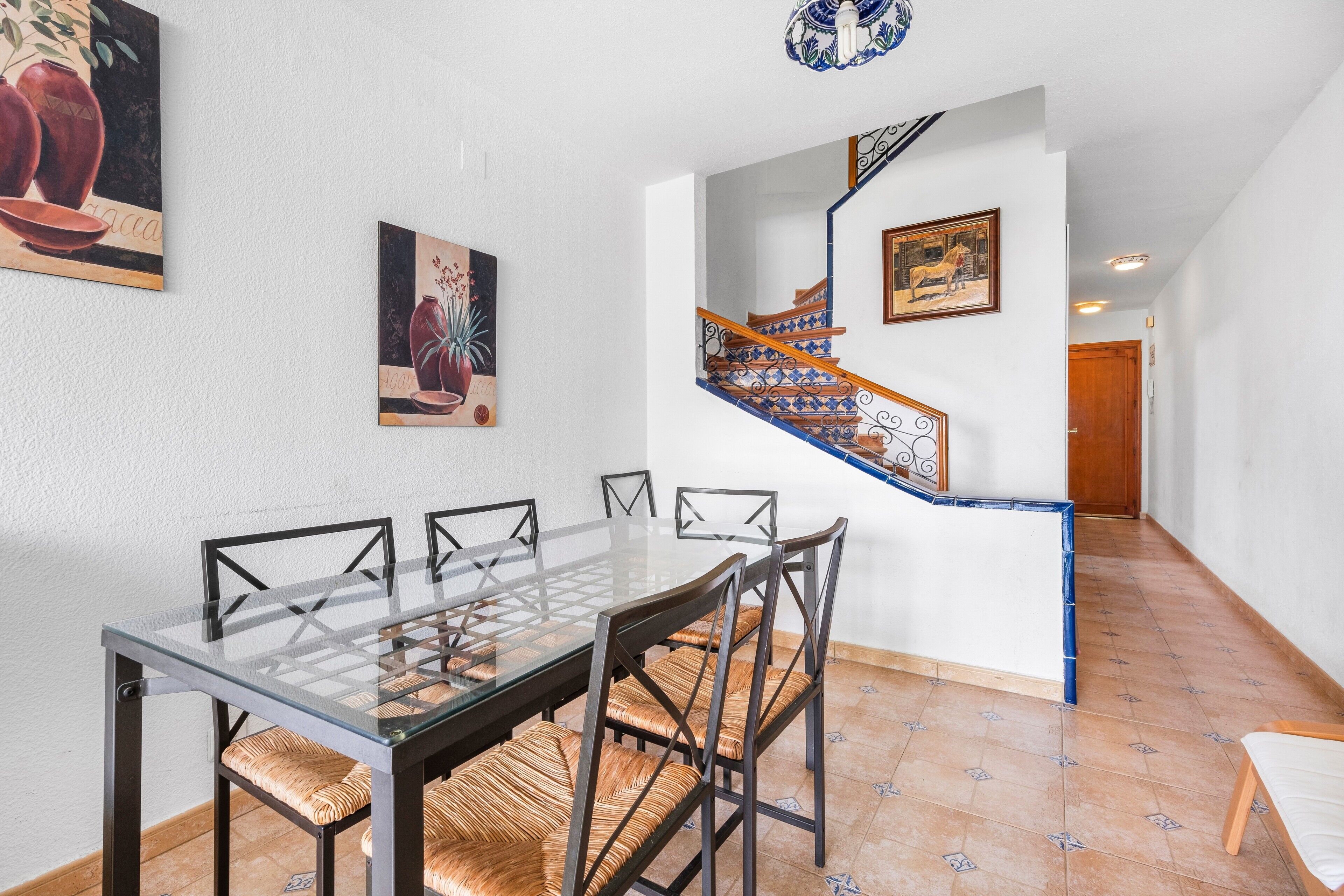 A dining area with a glass-top table, six chairs, and a staircase with blue and white tiles.