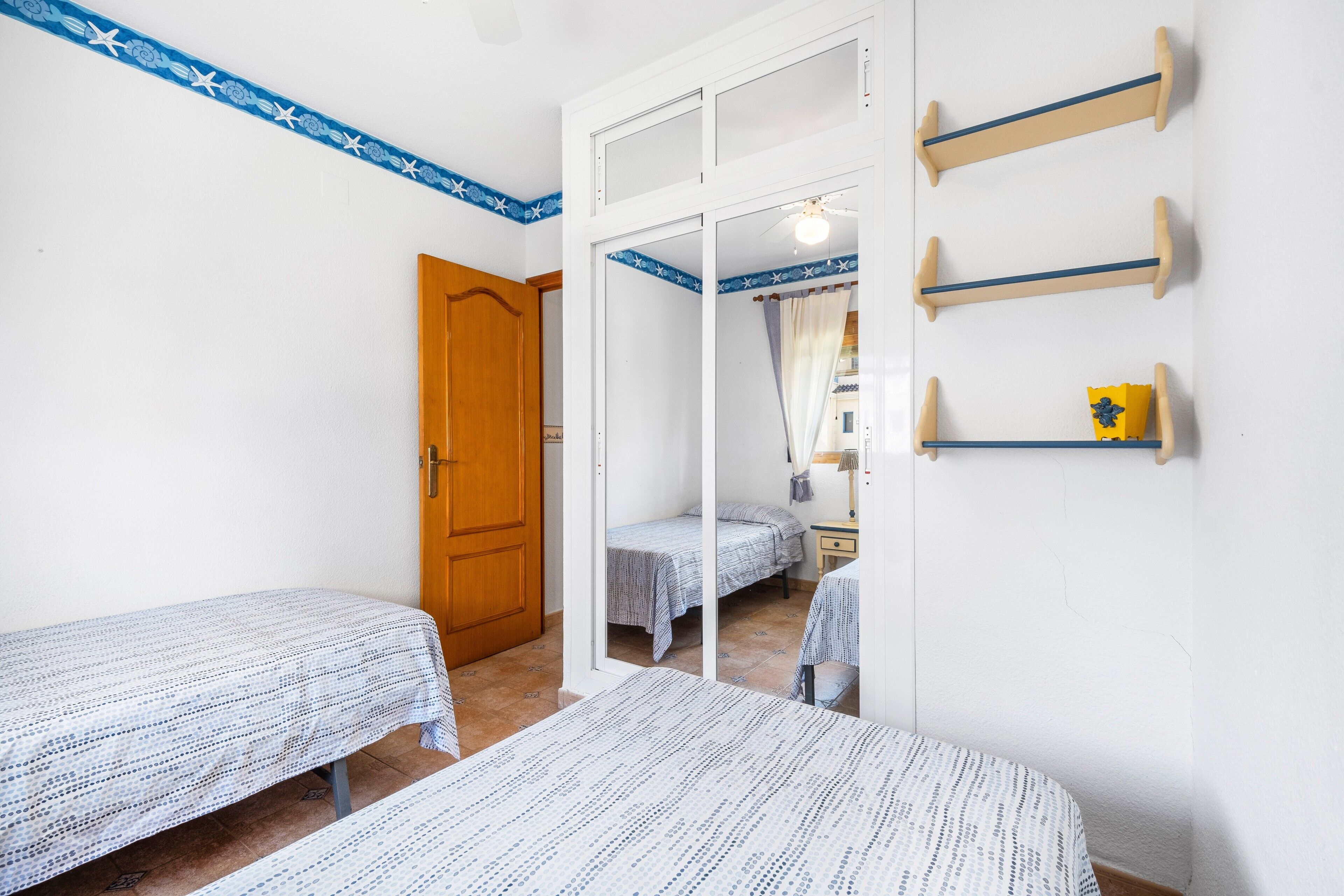 A room with two beds, a wooden door, a mirror, and a wall-mounted shelf with a small item.