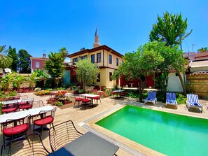 A pool area with lounge chairs and tables, surrounded by a garden with various plants and a building with a tower. - Necip Bey Konağı (Antalya)