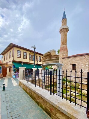 A historic building with a prominent minaret, a paved walkway, and a stone wall with a metal fence. - Necip Bey Konağı (Antalya)
