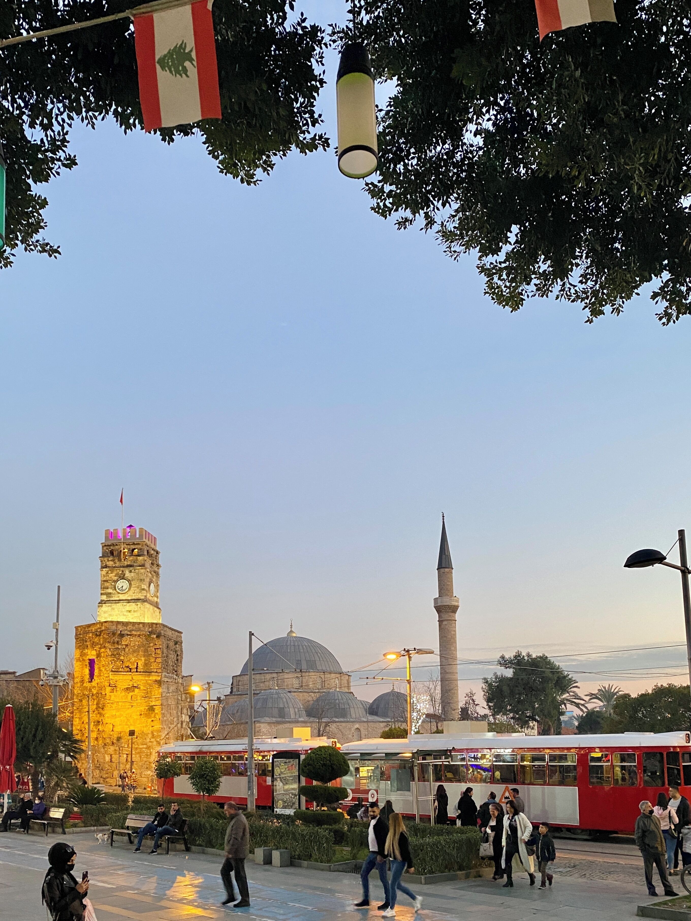 A public square with a historic tower, a mosque with a dome, and a tram.